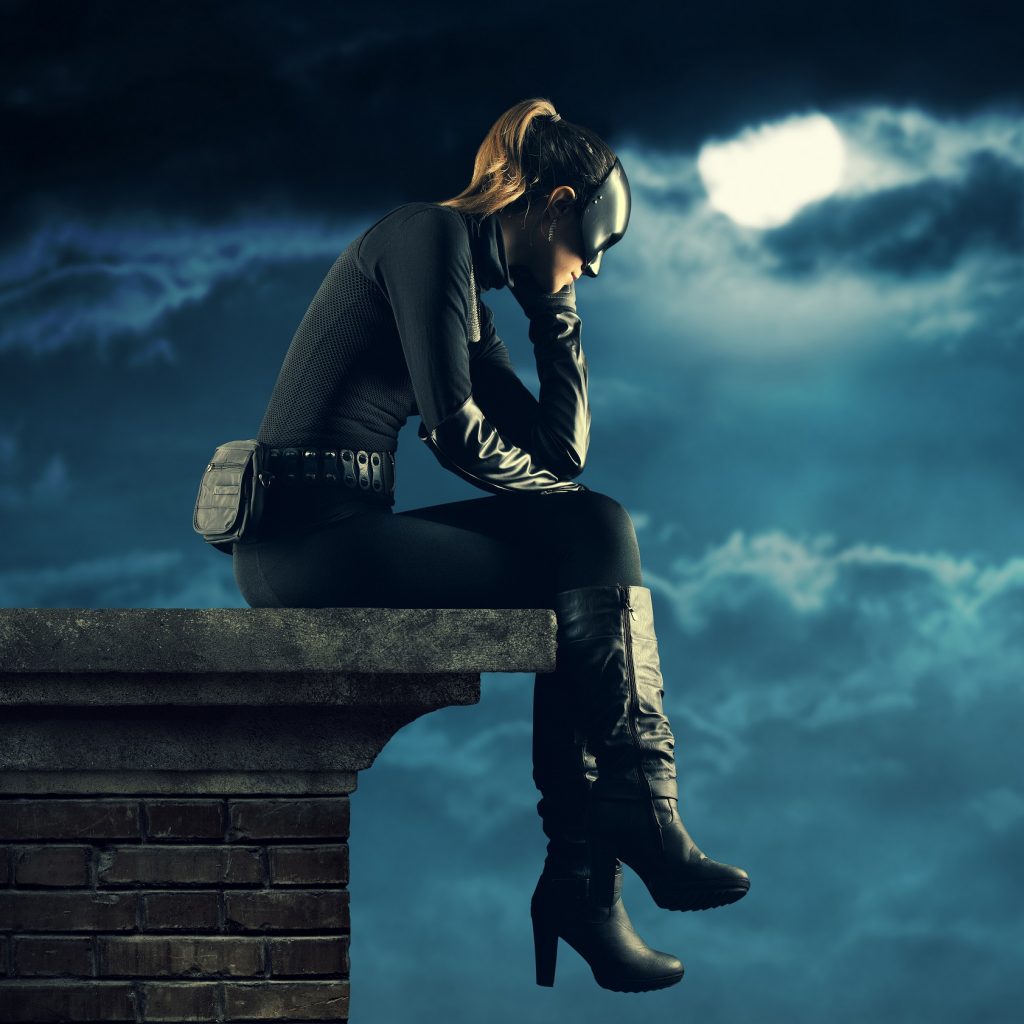 Woman in mask and catsuit, sitting and brooding on a rooftop against a cloudy moonlit sky
